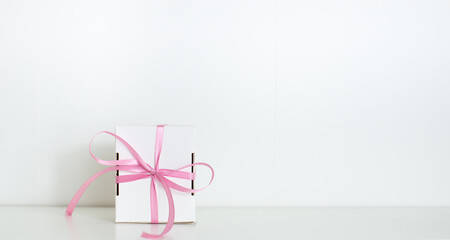 A  small gift with a pink ribbon on white background. White gifts with a pink bow. Christmas gifts. Gift for woman. Holiday pink background with gift, white satin bow, ribbon. 