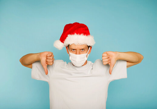 Young angry man wearing medical mask in red Santa Claus and white shirt showing hand ok sign down isolated on blue background.