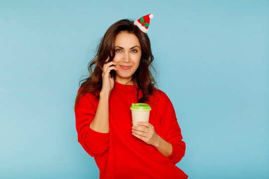Young beautiful santa claus woman talking by smartphone and drinking tea or coffee over blue background. Christmas and New Year lifestyle concept.