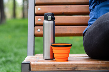 Steel thermos and silicone mug. Tourist dishes on the bench. Drinking coffee in the park.