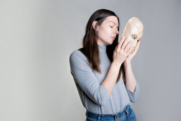 A young woman hides her face behind a mask, the concept of psychological problems, a crisis of self-identification or strong shyness. Photos in light gray tones, gray background.