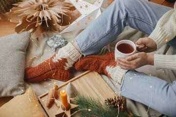 Christmas winter hygge. Woman hands with cup of tea, golden lights, christmas stars, pine, candle