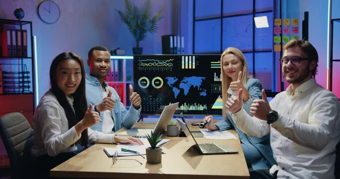 Close up portrait of likable smiling satisfied successful multiethnic workmates which gesturing thumbs up into camera while sitting behind the table in modern evening office