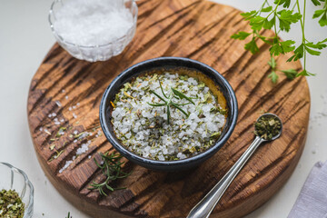 Fragrant seasoning, green salt with dried herbs in a ceramic bowl on a wooden board on a light background.
