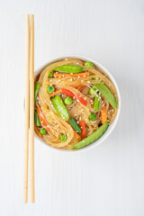 Top view of Korean Japchae salad made of stir-fried Cellophane noodles, green peas and beans seasoned with sesame seeds with savory sweet flavor served in bowl with chopsticks on white wooden table