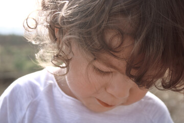 Niño pequeño de dos años con el pelo largo y rubio, a contraluz al aire libre con la cabeza agachada.