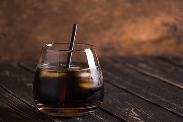 Beautiful glass with whiskey cola and ice with a black straw on a dark brown wooden table background. Delicious and elegant gossed alcoholic cocktail at the bar.