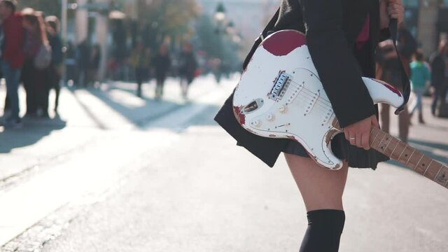 Hot woman walking with electric guitar. Daring rebel woman with purple hair playing electric guitar at city center during bright sunset. People walking in background. Asian girl like rock star