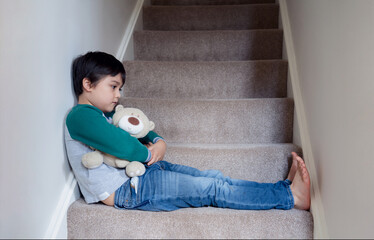 Sad asian boy sitting alone on staircase with teddy bear, Lonely kid looking dow with sad face not...