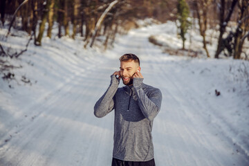 Happy sportsman putting earphones and listening to the music while standing in nature at snowy winter day. Technologies, winter fitness, healthy life