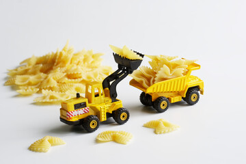 Toy yellow forklift truck with a bucket loads farfalle paste into the back of a heavy-duty dump truck. Concept of delivering food products, preparing for sale or cooking. White background
