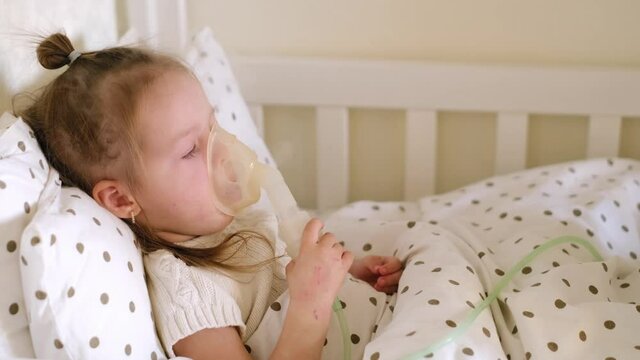 A sick child coughs violently during inhalation, inhaling vapors. Girl with an breathing mask is lying in bed under a blanket. Bed rest of a ill person. Respiratory diseases. Polka dot bed linen
