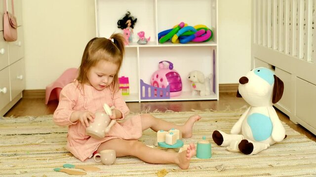 Girl is playing tea drinking, pouring a drink to her friend a toy soft puppy. Baby smiles amiably at the guest. Wooden set of toy dishes with treats. Kids games on the floor in the children's room
