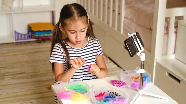 A cute little girl is recording an online lesson on weaving bracelets from rubber bands on a slingshot. A video on the topic of hand made is taken by a child on a smartphone for his blog