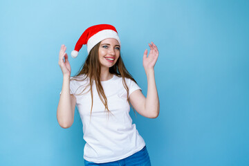Fototapeta na wymiar Portrait of woman in santa hat and white t-shirt posing playfully on blue background. Young caucasian woman joyful with anticipation of christmas discounts and gifts