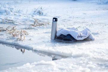 Thermos, slippers and a towel near the ice hole. Winter swimming. Hot drink in winter. Get warm with coffee. - obrazy, fototapety, plakaty