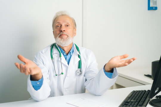 Portrait Of Mature Adult Male Doctor In White Uniform Coat With Stethoscope Speaking, Looking At Camera, Gesturing Spreads Arms Out To Sides, Sitting At Desk With Computer In Medical Office,