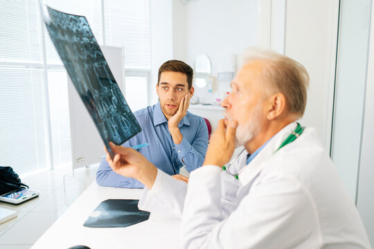 Pensive serious mature adult male physician consult shocked young man patient giving bad news explaining results of MRI image. Sad scared young man listening to bad news sitting in doctor office.
