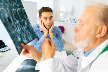 Obraz premium Close-up of mature adult male physician consult scared young man patient giving bad news explaining results of MRI image. Unhappy frustrated young man listening to bad news sitting in doctor office.