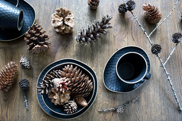 Dark blue set of dishes: tea cup, saucer, plate, a bowl with fir cones, variety of cones on wooden background. Christmas table decoration, magical holidays