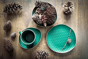 Emerald-colored set of dishes: tea cup, saucer, plate and variety of cones on wooden background. Christmas table decoration, magical holidays