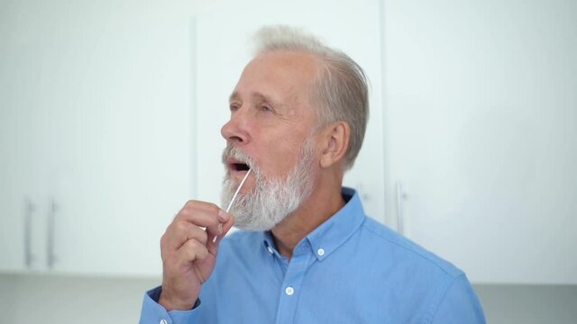 Close-up of mature Caucasian adult man taking swab sample of mouth carrying out coronavirus self home test. Handsome pensioner male taking swab probe from mouth with stick genetic paternity test.