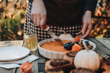 The woman cuts and serves the pie to guests. Thanksgiving festive table. Cozy autumn style table setting with pumpkins and leaves. Pumpkin pie and cinnamon rolls. Fall styled composition.