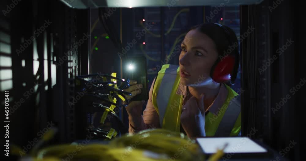 Poster caucasian female it technician wearing headphones and using torch checking computer server