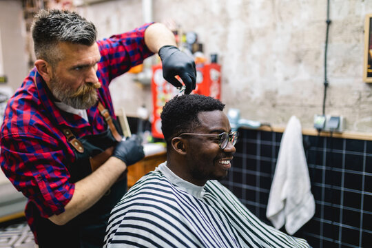 Young African Man Visiting Hairstylist In Barber Shop. Professional Hairdresser Cut Hair With Scissors.