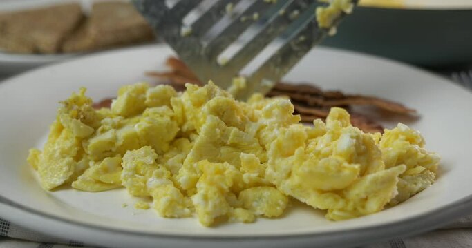 Spooning scrambled eggs onto a plate with bacon and toast slow motion