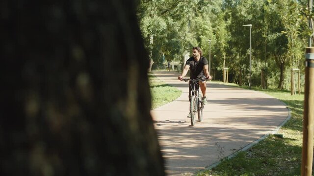 Long shot of man with bionic leg cycling along path in park. Tracking shot of cheerful bearded man riding bike in summer park, enjoying time outdoor, looking around. Disability, sport concept