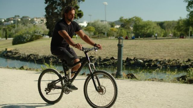 Long shot of focused man with prosthetic leg riding bike in park. Tracking shot of bearded Caucasian young man with bionic leg learning to ride bicycle alone in summer. Disability, sport concept