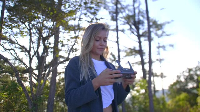 Blonde girl flying a drone, with a controller, in a mountain forest, on a sunny evening
