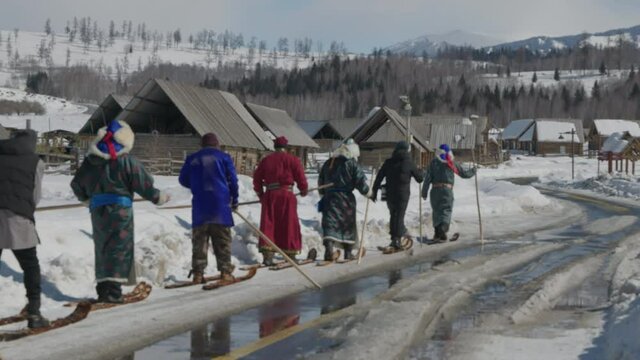 A group of men walking in ancient fur skis in single file line through Hemu Village, Xinjiang, China in traditional deel and fur ski on sunny winter day.