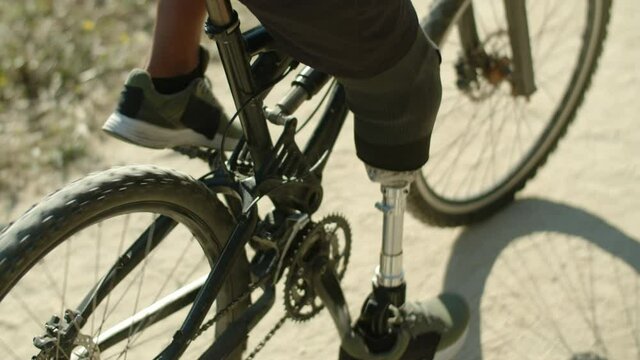 Close-up shot of man with artificial leg pedaling bike in nature. Back view of person with prosthetic leg spending time outdoors on summer day, riding bicycle along path. Disability, sport concept