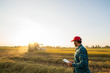 A smart farmer wearing red cap, jeans,shirt, operates tablet to control combine harvester in the...