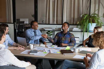 Happy middle aged african american leader shaking hands with biracial colleagues thanking for good ideas, praising for excellent work, having fun together at brainstorming meeting with diverse workers