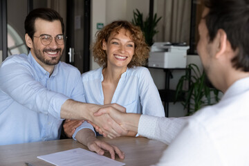 Joyful beautiful young family couple shaking hands with hispanic lawyer or broker, feeling thankful...