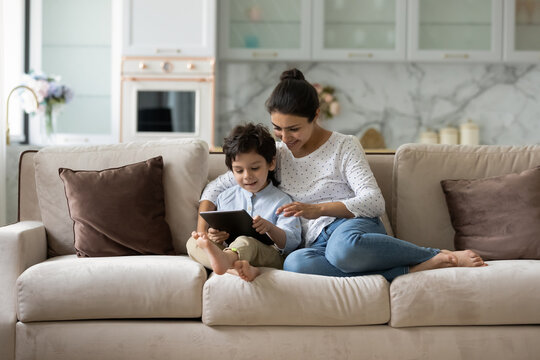 Happy Millennial Indian Mom Gen Z Son Boy Reading Book, Playing Videogame On Tablet Computer, Using Online App, Service, Chatting. Mother And Kid Shopping, Booking On Internet At Home