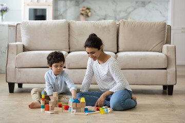 Fototapeta premium Happy young Indian mom helping preschool son to build toy towers from wooden construction blocks, playing with kid on heating floor at home. Babysitter watching child. Motherhood, daycare concept