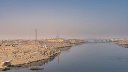 The Nile River in the area of the Aswan Dam. Technical buildings are visible on the sandy shores. Blue sky. Reflection on the smooth surface of the water. Egypt