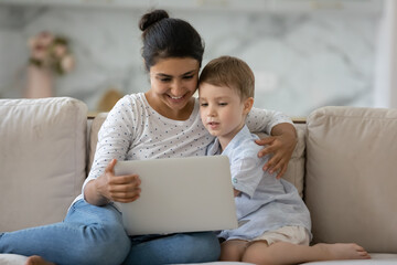 Curious kid and happy nanny holding laptop on sofa, watching online media content, playing games on internet together. Mom teaching son, using learning app on computer, reading book