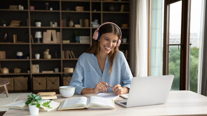 Happy attractive millennial woman in headphones watching interesting educational online lecture or holding video call with teacher learning foreign language, studying distantly using computer.