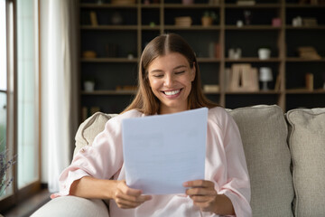 Happy young beautiful woman feeling pleased reading paper correspondence letter with good news, getting last bank mortgage payment notification or taxes refund, sitting on cozy couch at home.