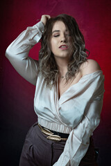 Super sexy, happy, fashionable and stylish girl plus size poses in a satin blouse with a deep neckline and bare shoulder, gray trousers and jewelry accessories on a red-pink gradient background 
