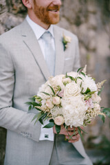 Groom with a bouquet of flowers stands against a stone wall