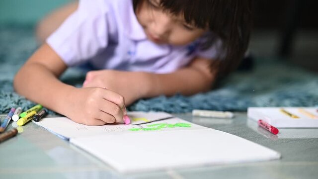 little girl writing and coloring with chalk crayon while relaxing on carpet at home after comeback from school