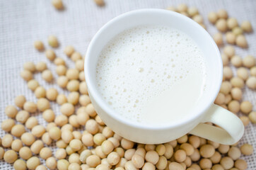 Top view of fresh delicious homemade soybean milk in soft yellow cup with dry soybean seeds on white sack fabric background