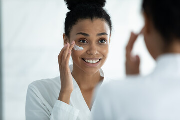 Happy beautiful young african ethnicity multiracial woman in bathrobe applying hydrating creme on face, looking in mirror, enjoying doing daily moisturizing skincare routine after showering at home.