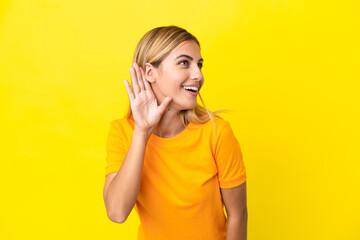 Blonde Uruguayan girl isolated on yellow background listening to something by putting hand on the...